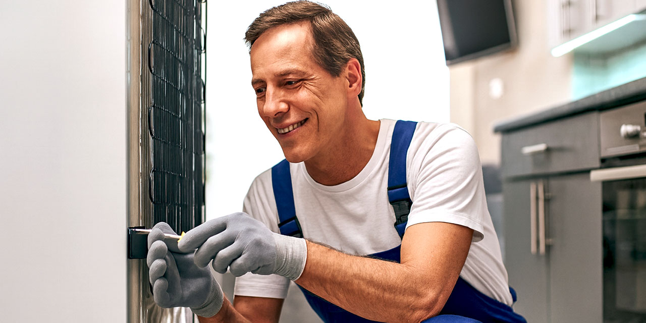 Smiling technician in blue overalls repairs the back of a refrigerator in a modern kitchen